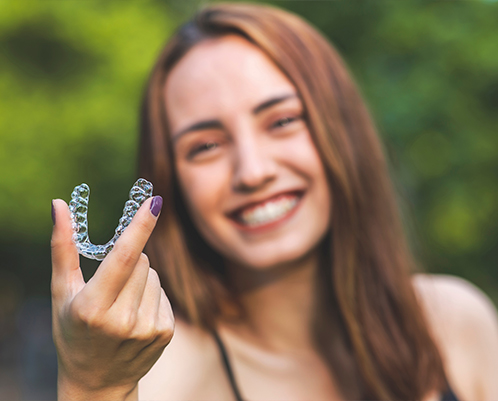 Smiling young woman holding Invisalign aligner outdoors with green trees in background