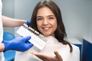 Woman with brown hair in dental chair gesturing to a shade guide being held to her teeth